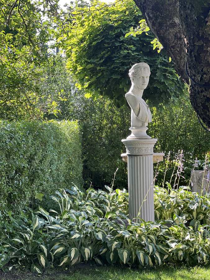 Statue på høy sokkel omgitt av hosta og klippet hekk